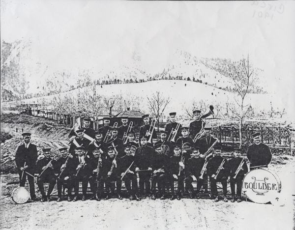 group of men in dark uniforms with caps, holding musical instruments. The bass drum head reads 'Boulder'. The Boulder Flatirons are in the background, possibly at Chautauqua Park.