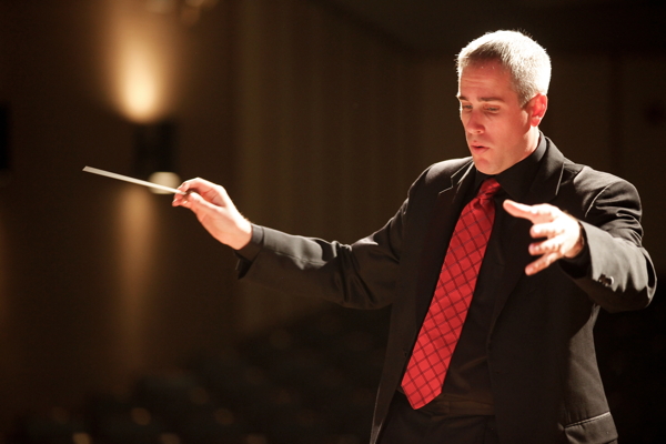 man in black suit, black shirt and red tie, conducting the band
