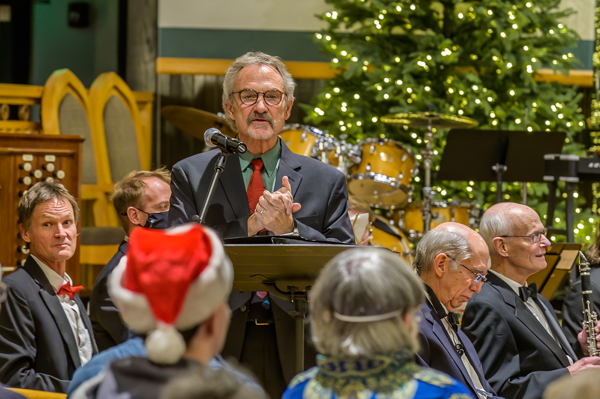 male narrator wearing red tie, surrounded by players and audience, in front of an evergreen tree with small white lights