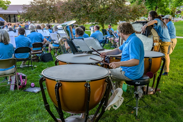 man in blue shirt playing timpani, with other players seated in front of him and two players standing to his right