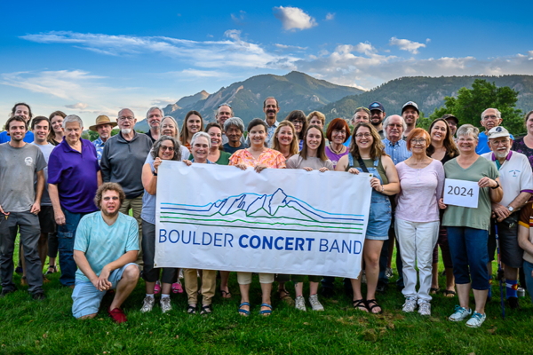 group of smiling people in summer clothing, holding a banner for the Boulder Concert Band. The Boulder Flatirons are in the background, beneath a blue sky