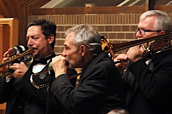 three men dressed in black attire. One plays trumpet, one plays trombone and the third man plays harmonica