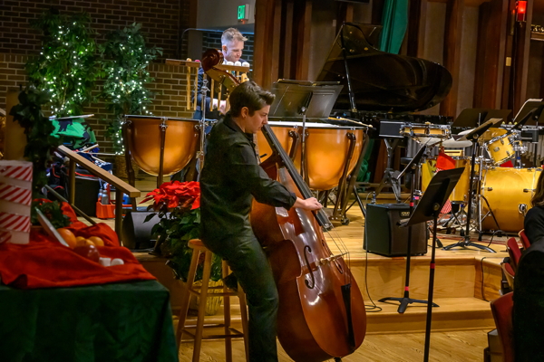 person dressed in all-black, seated on a tall stool, plucking an upright string bass