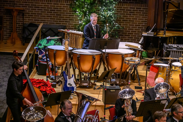 man in a black suit and white shirt, playing four timpani. A string bass player and a few low brass players in the foreground.