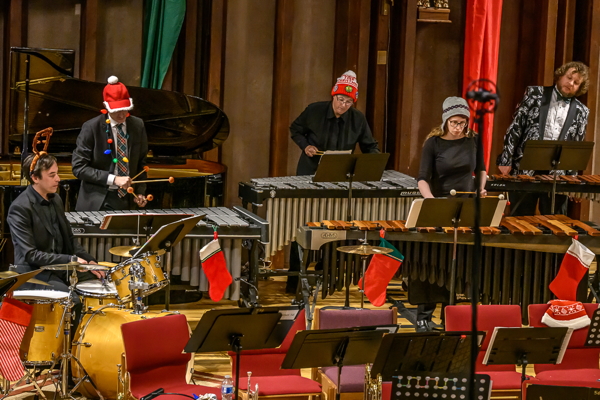 several people dressed in Christmas attire, playing keyboard mallet instruments, one playing a drum set. One of the players is wearing a string of Christmas lights around his neck.