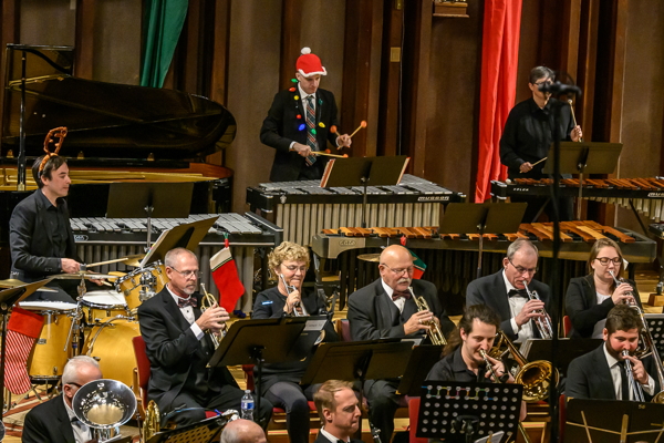 men and women seated and playing trumpets and trombones, behind them a player on drum set and two people playing mallet instruments