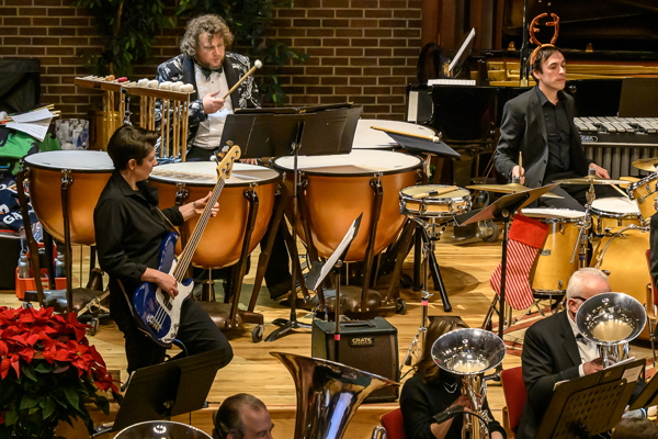 three people dressed in balck, one playing electric bass, one at a maple wooden drum set, and one playing four timpani