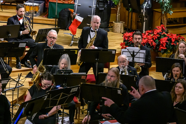 man in black tux, white shirt, black bow tie, standing and playing the tenor saxophone, surrounded by other players in the band. There is a group of red poinsettias in the background.