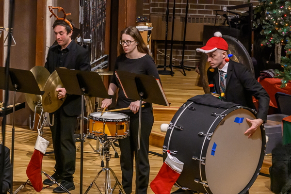 three people dressed in black. One is playing cymbals, one the snare drum, and the third is playing bass drum. He is wearing a red baseball cap with white fur trim