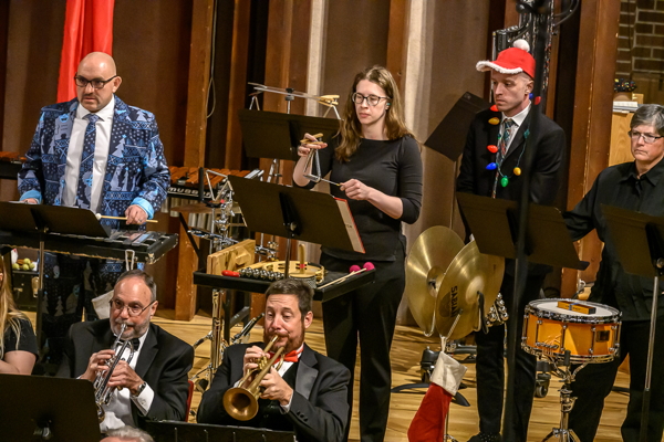four people playing percussion -- bells, triangle, cymbals and snare drum. Two trumpet players are seated in front of them.