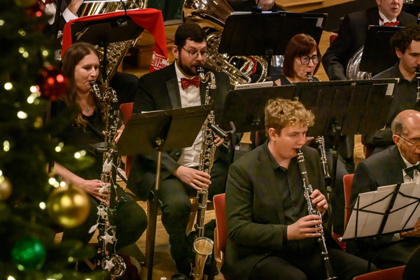 people seated in black attire, one with a red bow tie, playing bass and soprano clarinets. There is a decorated Christmas tree to the left