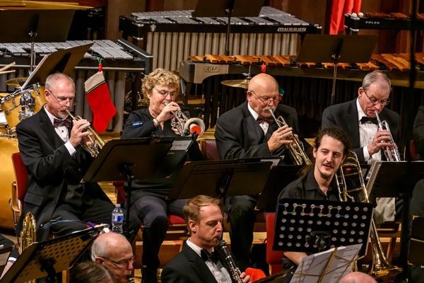 four people seated and playing trumpet, with a few other players seated in front of them