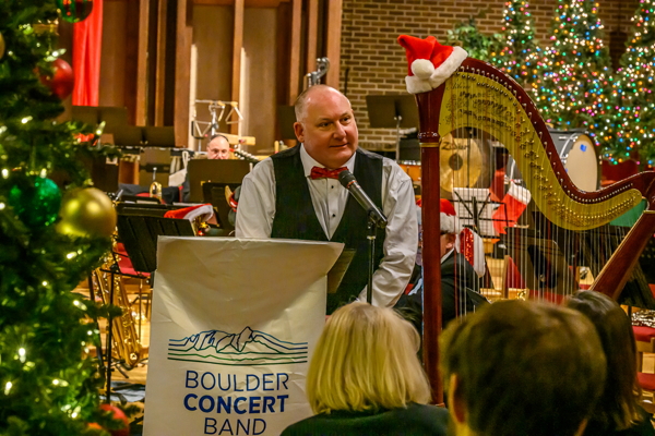 man in white shirt, red bow tie and black vest, standing next to a harp with a Santa hat on it