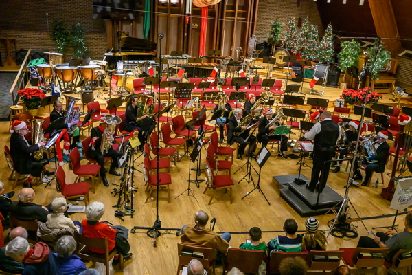 people seated in a semicircle, playing tubas and euphoniums, facing a man with white shirt, black vest and black pants