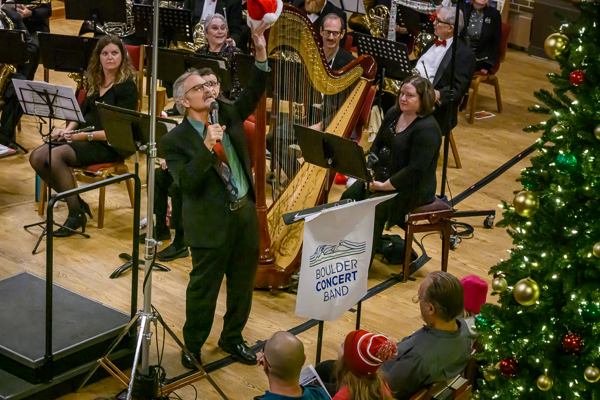 man in blue shirt, long tie and black suit, standing behind a music stand bearing the Boulder Concert Band banner