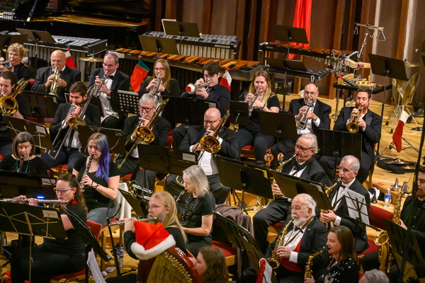 men and women in black concert attire, playing clarinets, flutes, saxophones, trombones, and trumpets. In front is a harp with a red and white Santa hat perched on top