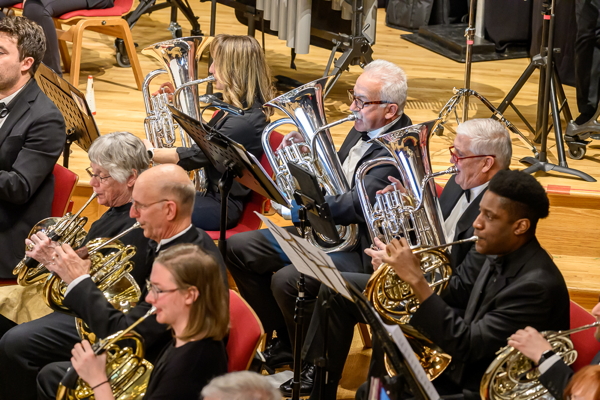 seven people seated in all black attire, playing french horns and euphoniums