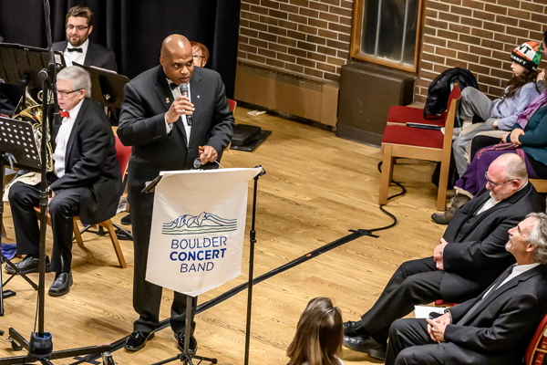 man in black tux, speaking into a microphone behind a banner reading Boulder Concert Band. There are men seated and facing him.