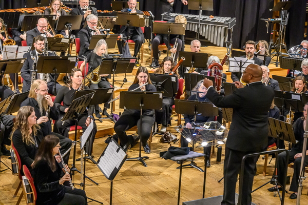 man in black suit, conducting a group of people in black and playing wind instruments