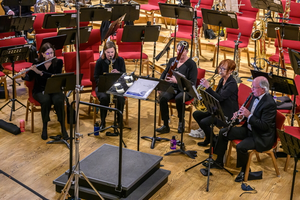 five people seated in front of a group of empty chairs, playing flute, soprano saxophone, bassoon, french horn and clarinet