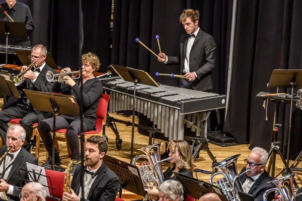 people dressed in concert black attire: two men playing saxophones, and man and a woman playing trumpets, a man standing and playing a vibraphone with four mallets, and a woman and a man playing euphoniums