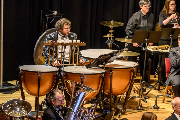 man with curly hair and glasses, wearing a black and white formal jacket, is playing four timpani