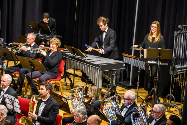 man playing vibraphone, flanked by a man playing orchestra bells and a woman using mallets on a suspended cymbal. Several wind players are seated in front of them.