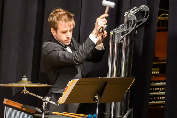 man dressed in black with a white shirt, using mallets to play the chimes. He is looking at his music on a black music stand. A suspended cymbal is beside him, and a chime tree is below the cymbal.