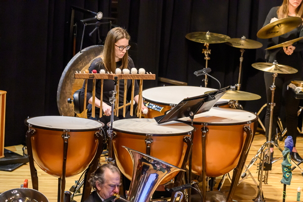 woman wearing glasses, using mallets to play four timpani. Several suspended cymbals are arrayed to one side.